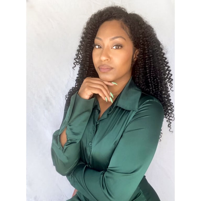 Woman with long curly black hair wearing a dark green button-up shirt, posing with hand near face against a white background